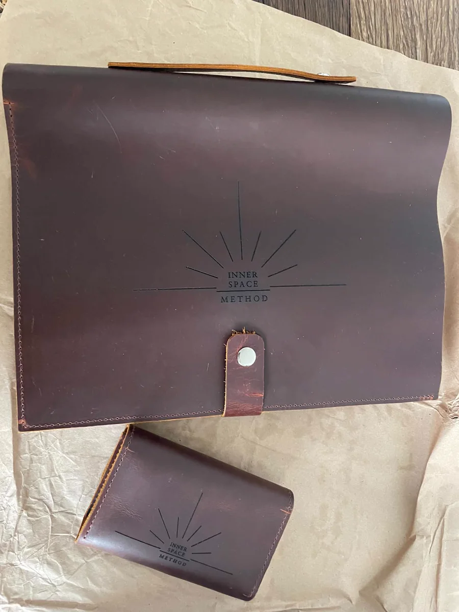 The finished mark, embossed in leather: the radiating Inner Space Method logo on the practice folio, a smaller card wallet beside it. Das fertige Zeichen, in Leder geprägt: das strahlende Inner-Space-Method-Logo auf der Arbeitsmappe, daneben ein kleines Kartenetui.
