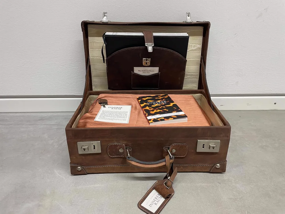The travelling installation: an open leather suitcase holding the book INNERER RAUM, the five phase workbooks and a kraft card asking what remains of space without place. Die reisende Installation: ein offener Lederkoffer mit dem Buch INNERER RAUM, den fünf Phasen-Workbooks und einer Kraftpapier-Karte mit der Frage, was von Raum bleibt, wenn der Ort verschwindet.